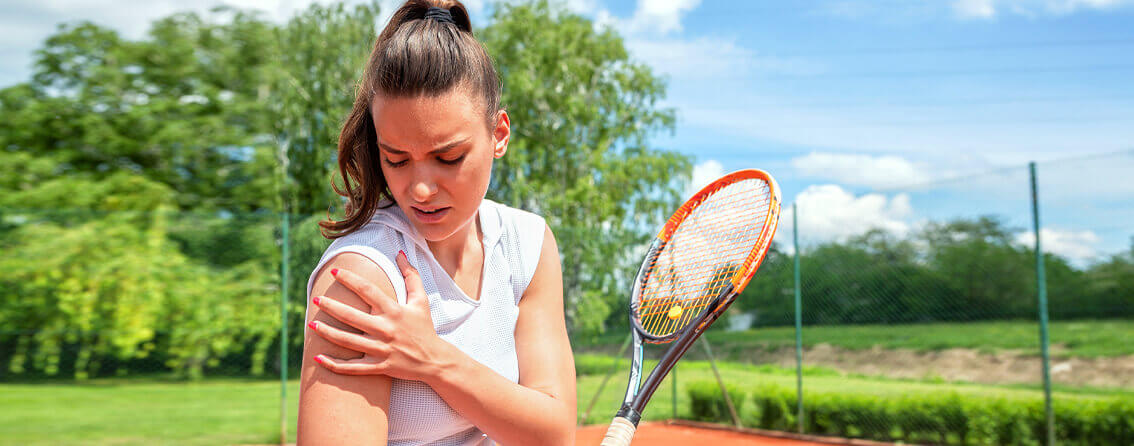 Tennis player holding a tennis racket in one hand and holding her shoulder in the other. She looks like she is suffering from shoulder pain.